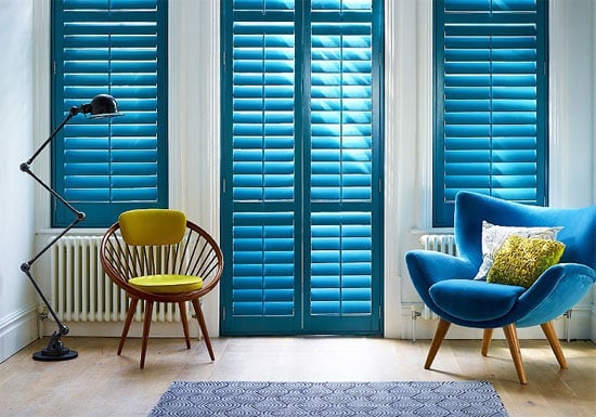 A vibrant living room featuring teal interior shutters on two windows and a central glass door