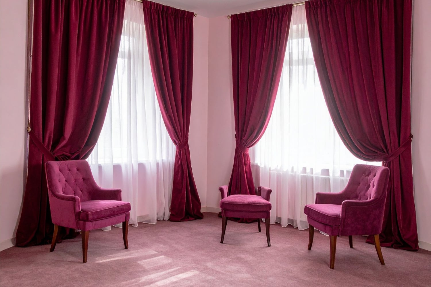 Blog » An elegant room features two windows framed by heavy burgundy velvet curtains and white sheers, with two matching tufted armchairs and a stool on a pink carpeted floor.