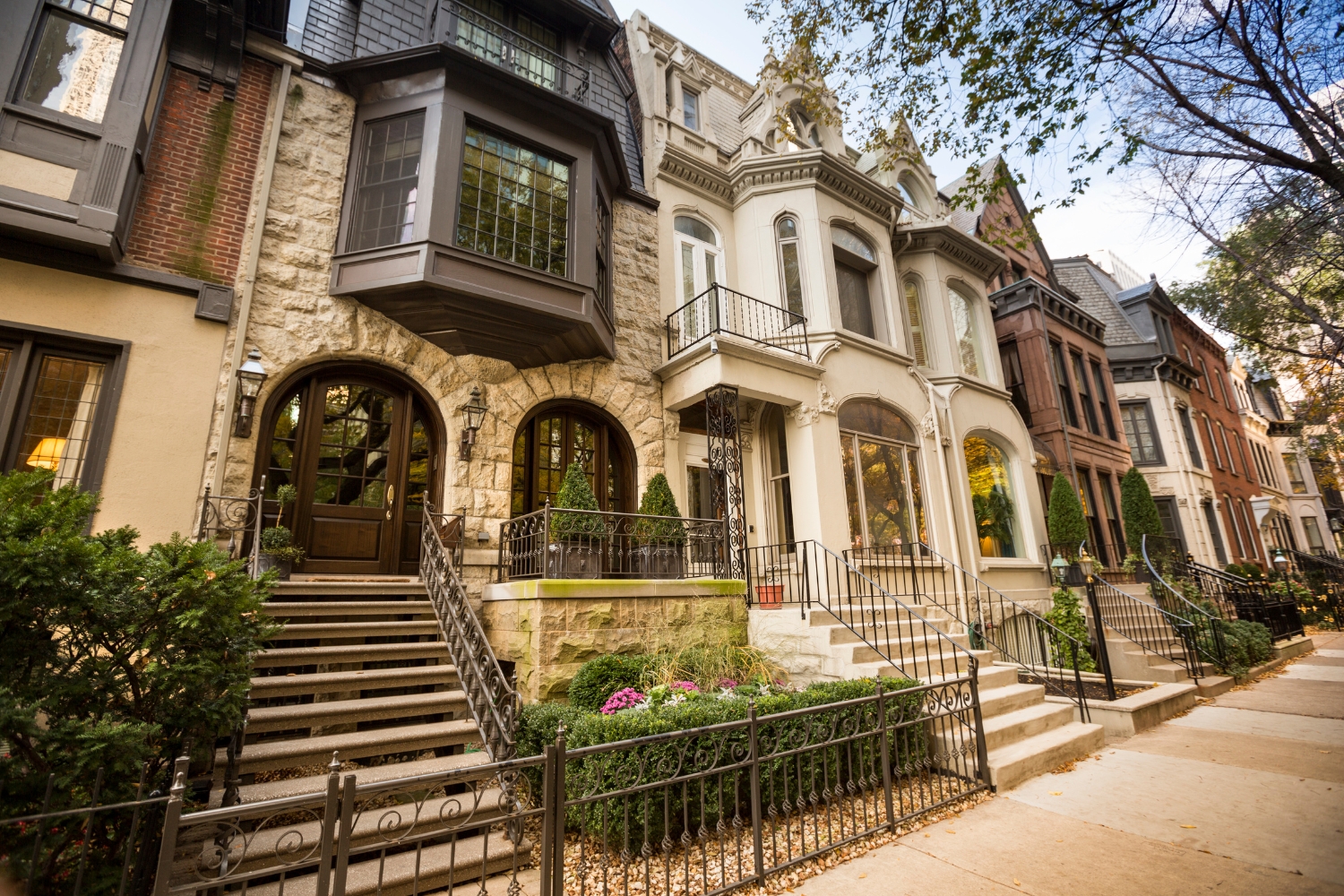 Blog » A row of historic stone and brick brownstone houses features ornate architectural details, arched entryways, and small front gardens behind iron fences.