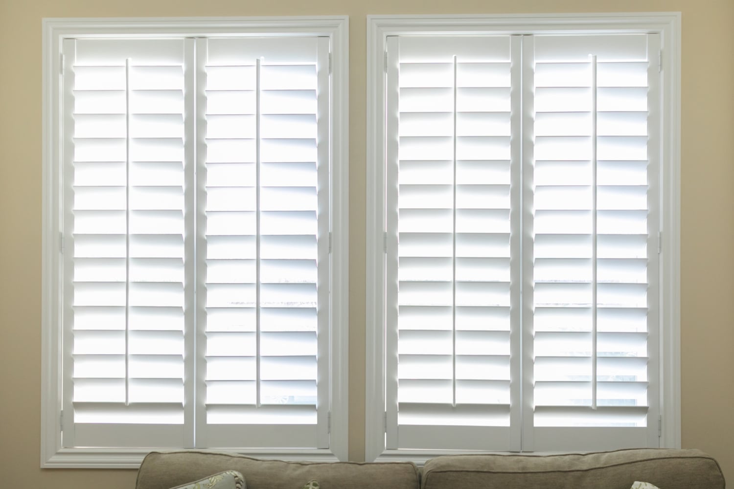 Blog » Two identical white plantation shutters are closed over two side-by-side windows above the back of a neutral-coloured sofa.